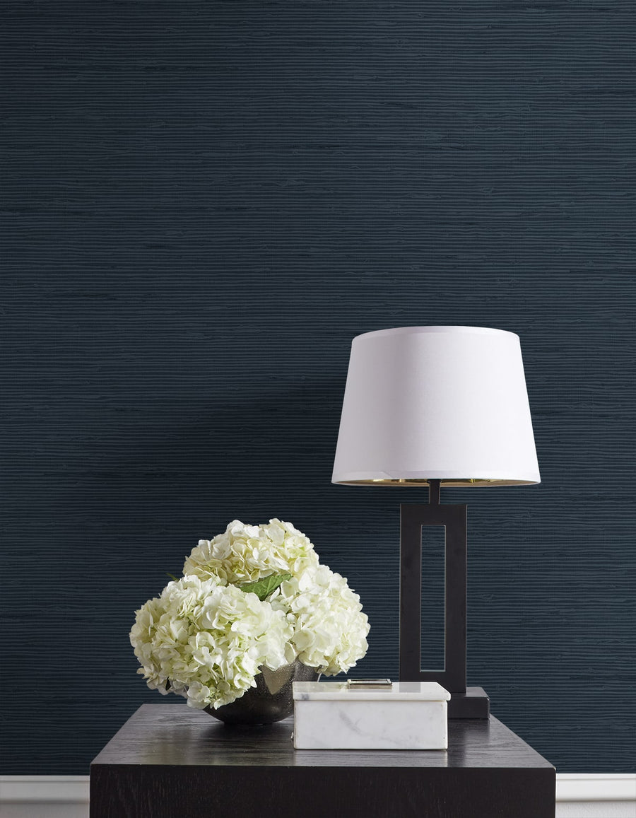 White lamp and floral arrangement on a dark wooden table against a dark blue textured wall.