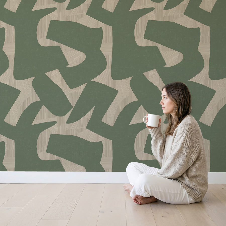 Woman sitting on the floor holding a mug in front of a Abstract Intarsia Wallpaper – Sage Green & Cream Textured
