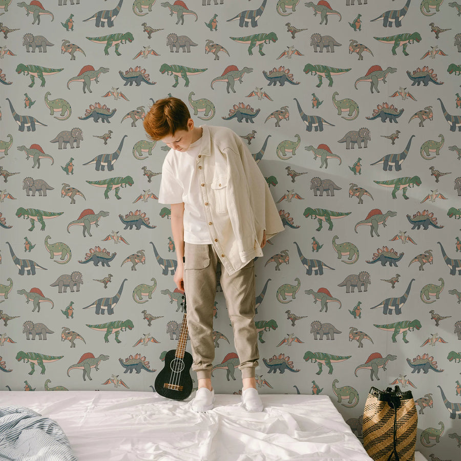 Sage green cartoon dinosaur kids wallpaper on a full wall behind a boy standing on a bed with a ukulele