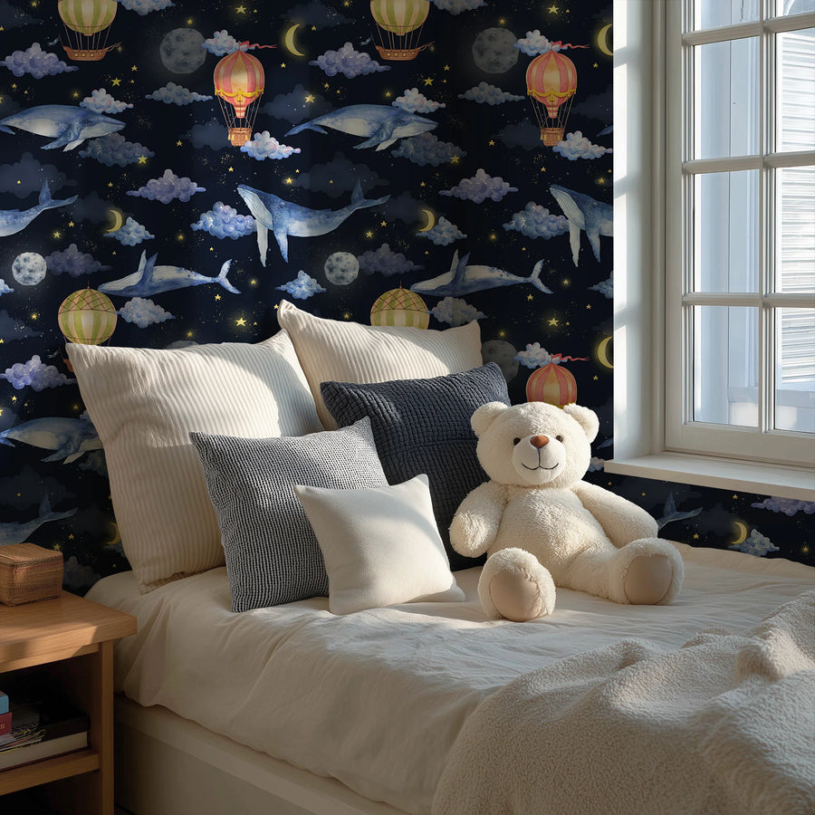 Hot air balloon and whale nursery wallpaper on midnight navy behind a bed with cushions and a teddy bear